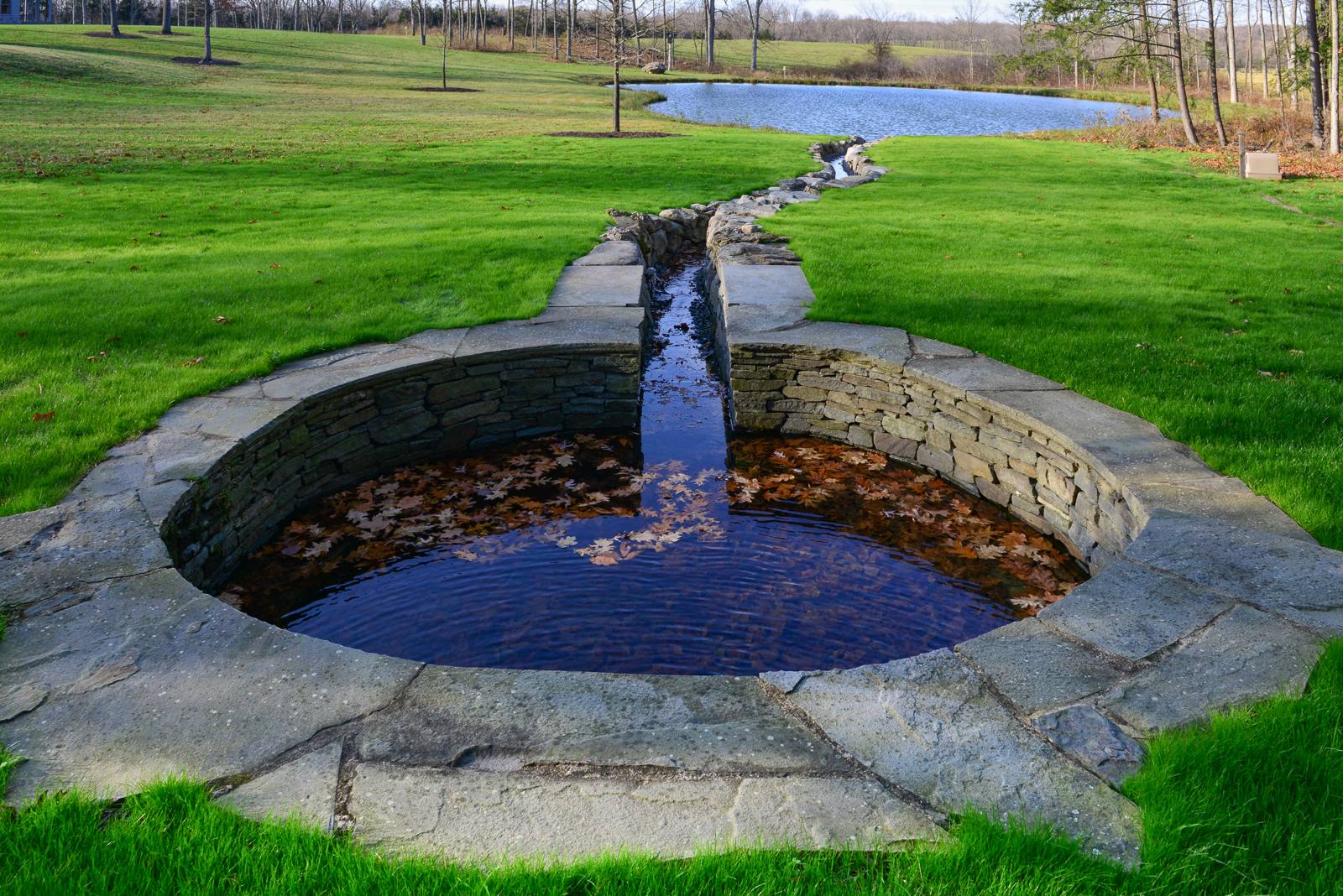 Stone-lined channel flowing from a round stone pool toward a pond