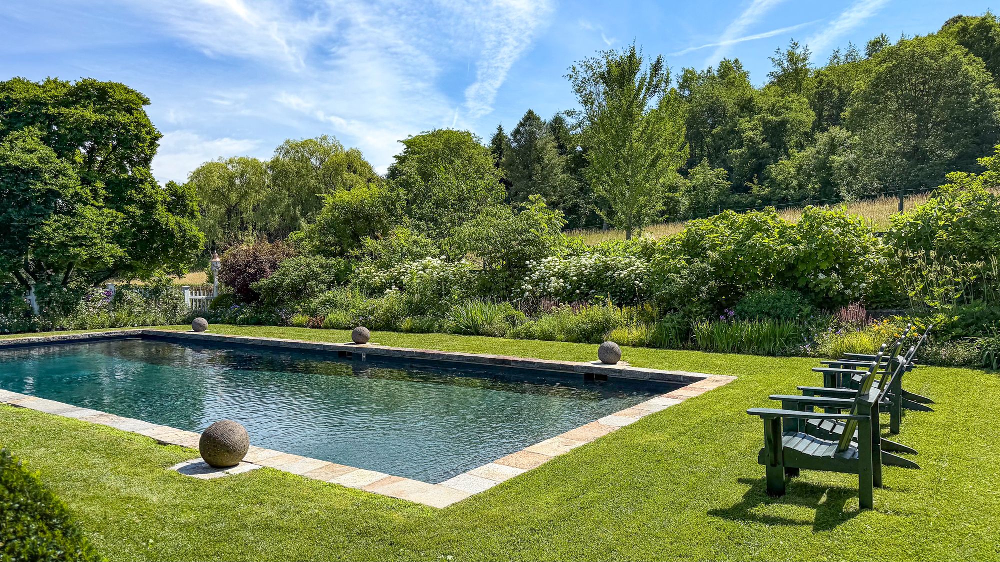 Stone-edged swimming pool set in a lawn with stone spheres, green Adirondack chairs and deep flowering borders